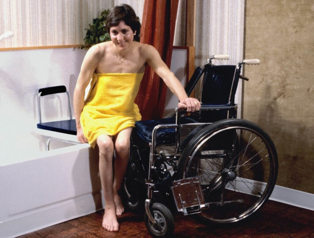 A woman in a yellow towel sits in a wheelchair next to a bathtub, smiling slightly as she reaches towards the tub, demonstrating the practicality of NDIS-approved housing modifications.