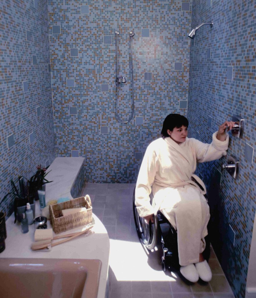 A person in a wheelchair reaching for the shower controls in a Specialist Disability Accommodation bathroom with blue mosaic tiles.
