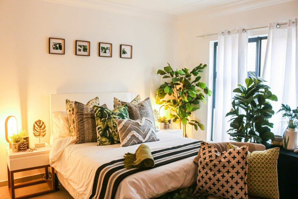 A cozy bedroom with a bed adorned with patterned pillows, framed plant pictures on the wall, large potted plants suitable for NDIS housing, and a bright window with white curtains.