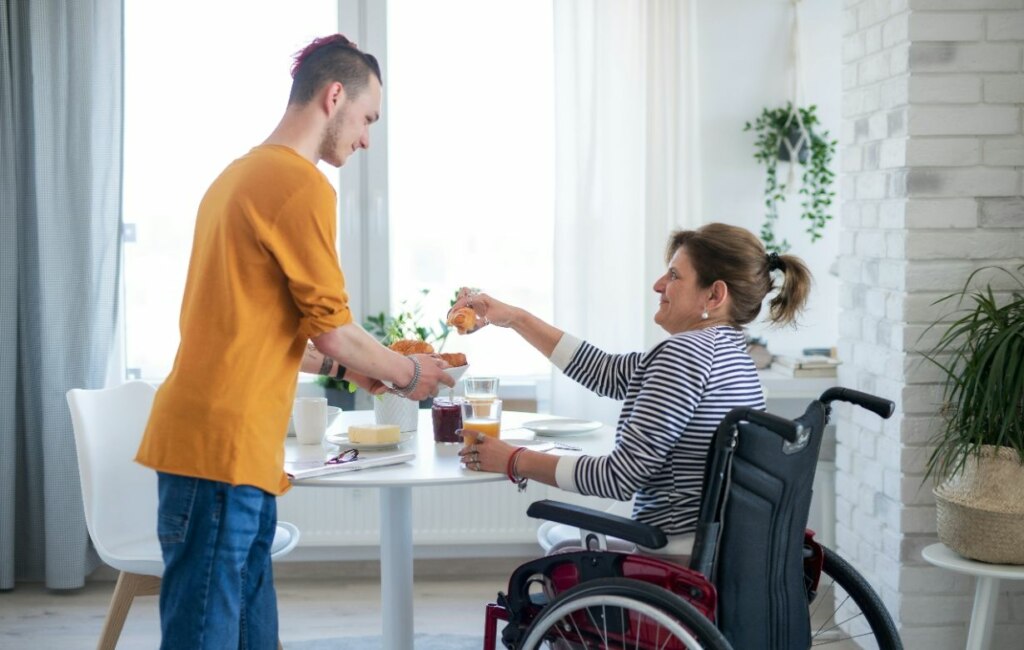 A young man serving breakfast to an older woman with disability support in a wheelchair at a home dining table.