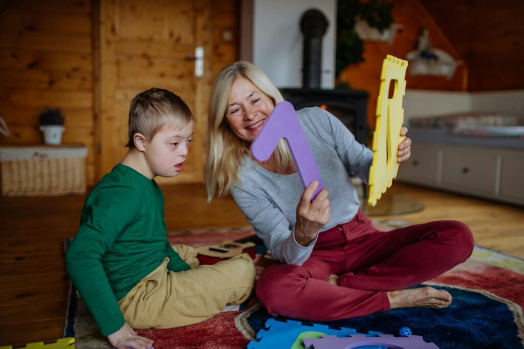 A woman and a young boy with Down syndrome, supported by the NDIS, sitting on a rug and playing with large letter-shaped puzzles in a cozy wooden room.