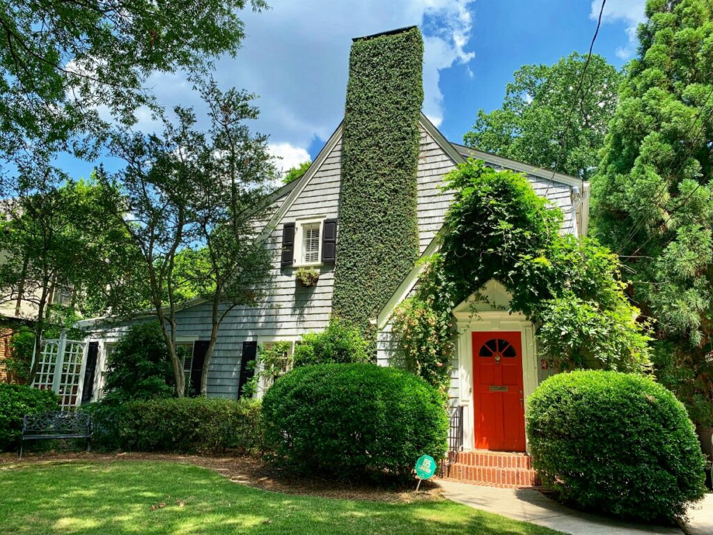 A charming suburban house with a gray facade partially covered in ivy, featuring a bright red door and surrounded by lush greenery under a clear blue sky.