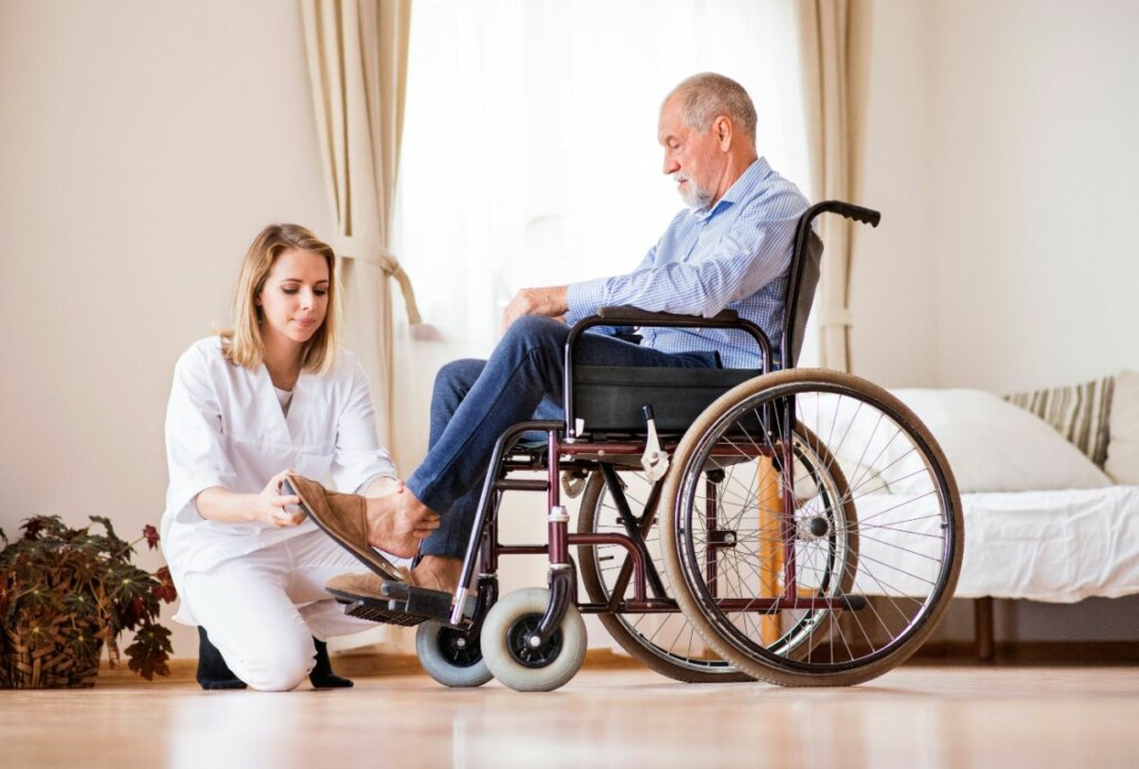 A healthcare worker kneels beside an elderly man in a wheelchair, both looking at a document she is holding in a bright room.