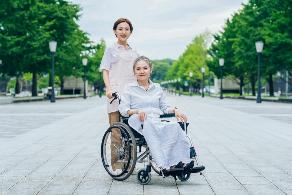 A caregiver stands behind an elderly woman in a wheelchair, both smiling in a tree-lined park walkway.