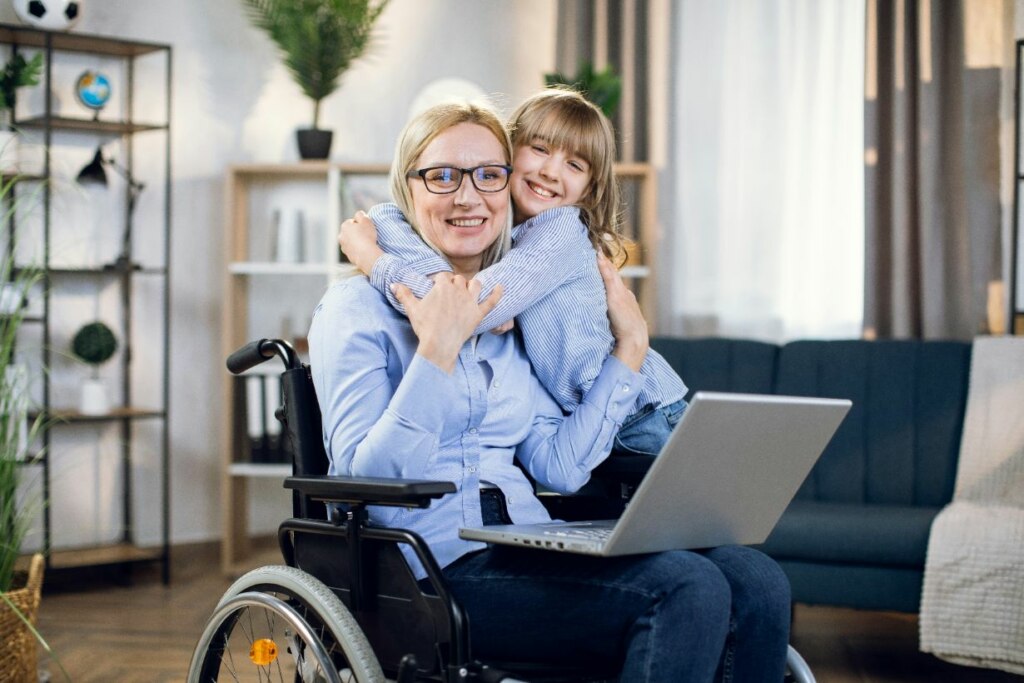 A smiling woman in a wheelchair hugs a young girl in a living room, with an NDIS application open on the laptop in front of them.