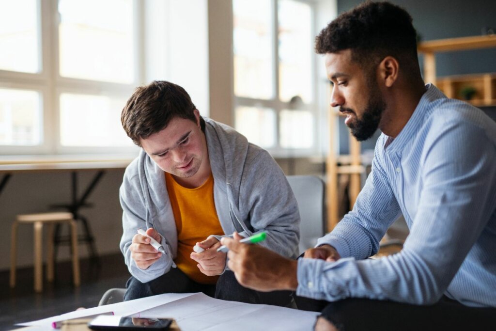 Two men discussing NDIS documents at a table, one holding a pen, in a brightly lit office environment.