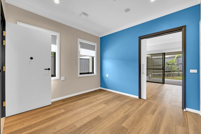 A bright room with light wood flooring, beige and blue walls. It features two windows, a white door, and sliding glass doors leading to an outdoor area. White ceiling lights illuminate the space, reminiscent of the airy homes in the Pendle Hill area of Sydney.