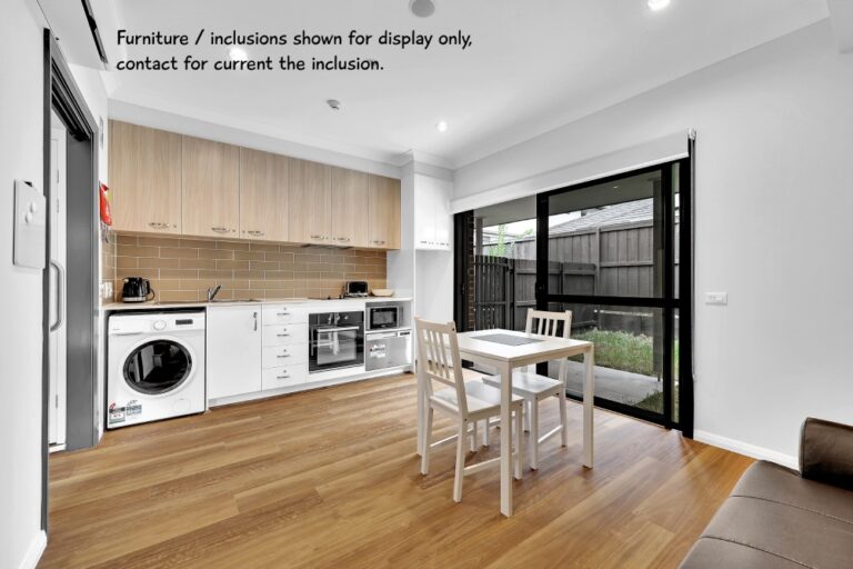 A modern kitchen with wooden cabinets, a washing machine, and an oven in the heart of Sydney. A small dining table with two chairs sits in front of glass doors leading to an outdoor area. Text indicates furniture is for display.