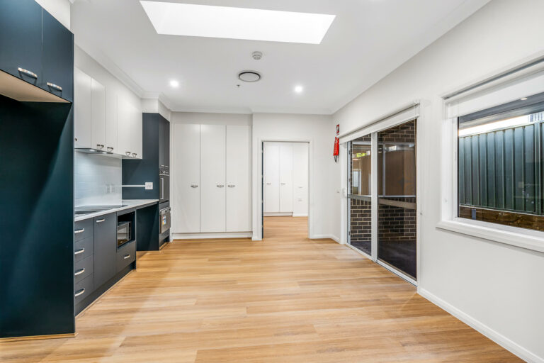 A modern kitchen in Wamberal with wooden floors, dark cabinets, and white countertops. A window lets in natural light, complemented by a skylight and multiple doors. Located on the picturesque Central Coast of NSW.