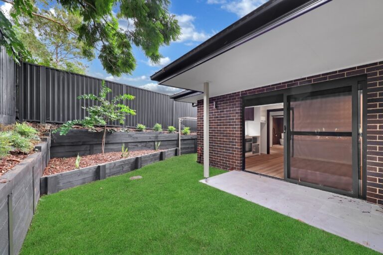 A small backyard in Concord boasts a covered patio, green lawn, brick wall, and raised garden beds filled with plants. A sliding door leads to the interior of the house in this charming Inner West Sydney residence.