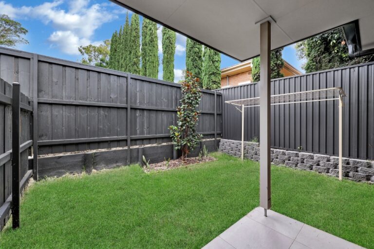 A small backyard in Pendle Hill with green grass, a few trees, a black wooden fence, a clothesline, and a covered patio area. The sky is partly cloudy.