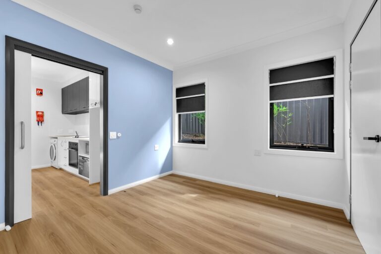 A small, unfurnished room with wooden flooring. The doorway leads to a compact kitchen with a washing machine, cabinets, and appliances. Two windows have blackout roller blinds. The white and blue walls add a touch of serenity. Conveniently located in Pendle Hill, near Parramatta in Sydney’s vibrant district.