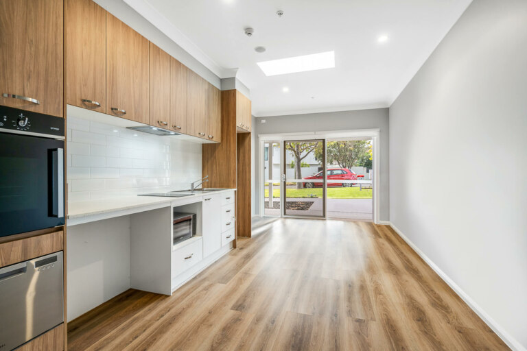 A modern kitchen with wooden cabinets, stainless steel appliances, and a white countertop opens into a bright living area with large windows overlooking the vibrant streets of Inner South Sydney, where a red car is parked outside.
