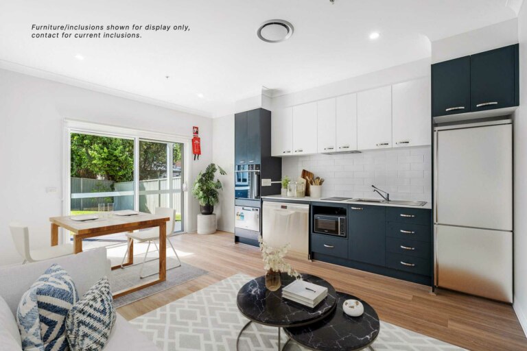 Modern kitchen and dining area with white and dark blue cabinets, stainless steel appliances, a wooden dining table set near a large window, and a cozy living area with a patterned rug and coffee table. Located in the heart of Sydney's Sutherland Shire at 30A.