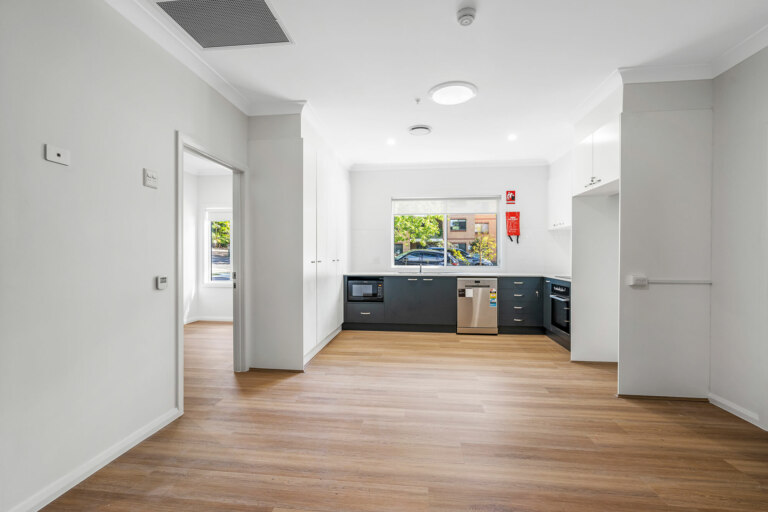 A modern, empty kitchen in Sutherland Shire, Sydney features wooden floors, white walls, and grey cabinets. Equipped with a microwave, oven, and dishwasher, it boasts a large window with an outside view. A doorway leads to another room.