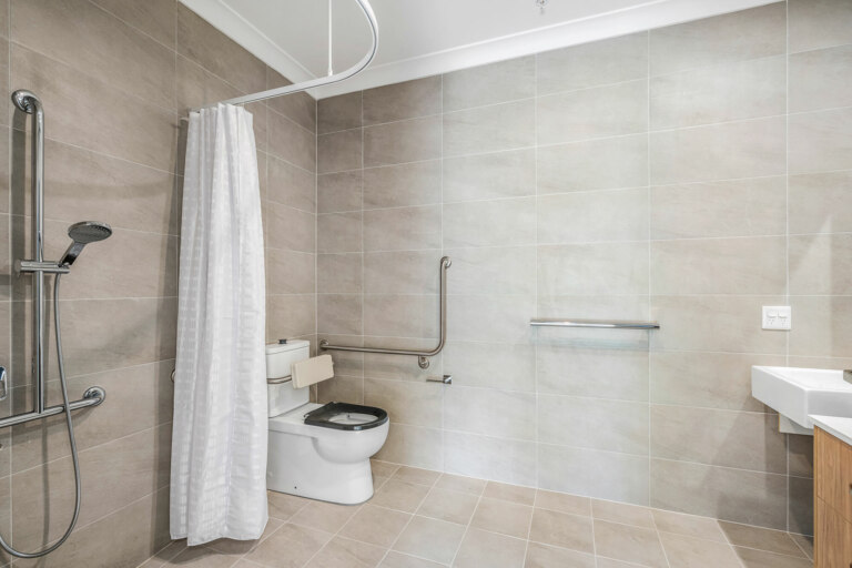 A modern bathroom in Sydney's Sutherland Shire features beige tiles, a shower area with a curtain, a wall-mounted handrail, a toilet with a side support rail, and a pristine white sink.